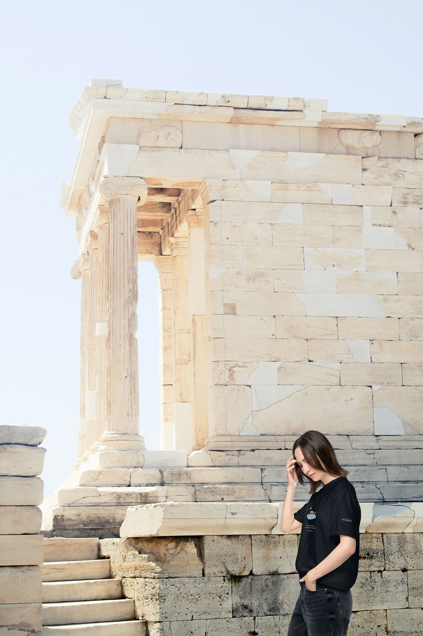 Young woman wearing a black unisex NOMADkit T-shirt with Acropolis Parthenon design, walking near ancient columns in Athens.