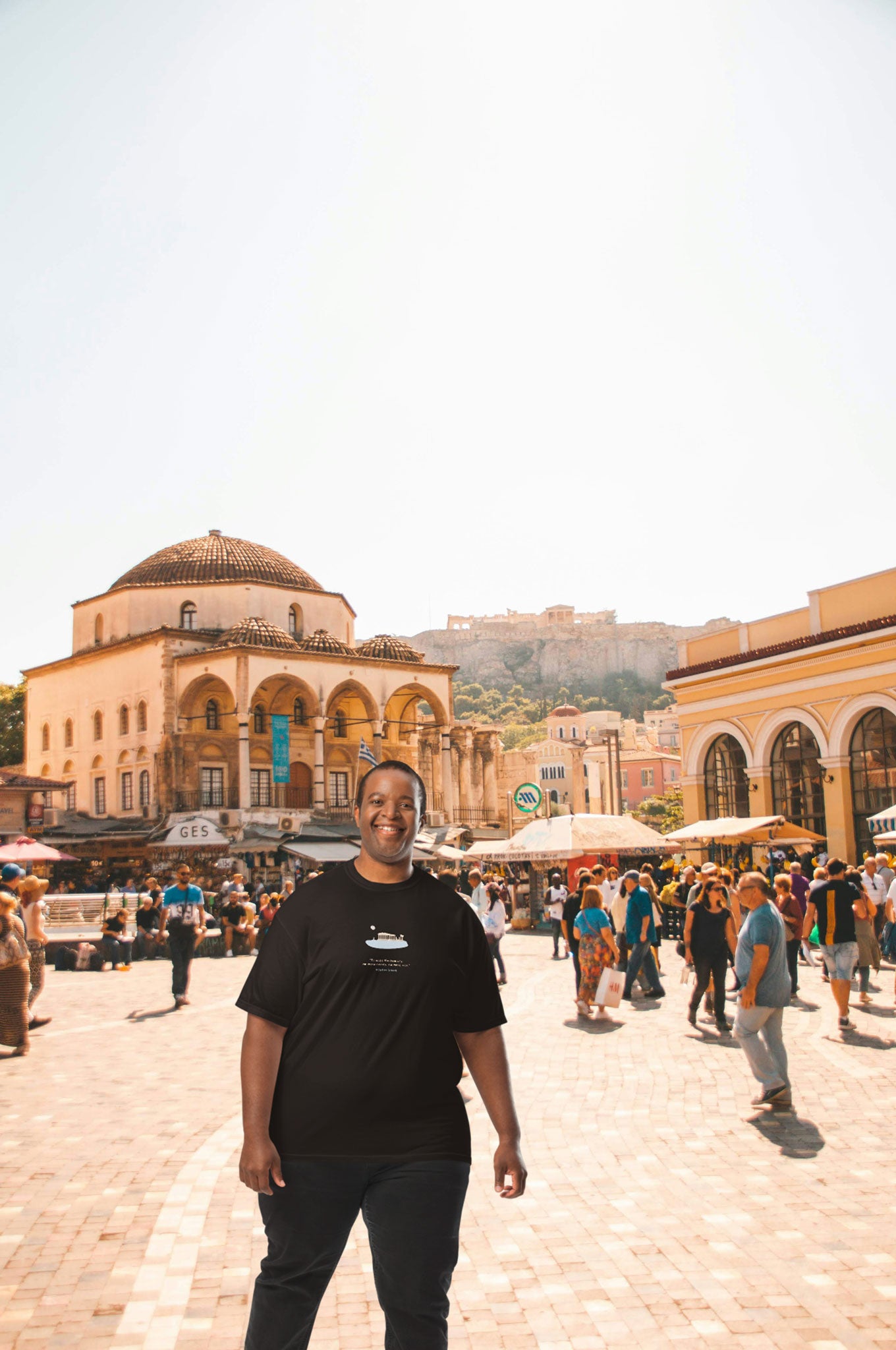 Man in black unisex NOMADkit T-shirt with Athens Parthenon design, standing in Monastiraki Square, Athens.
