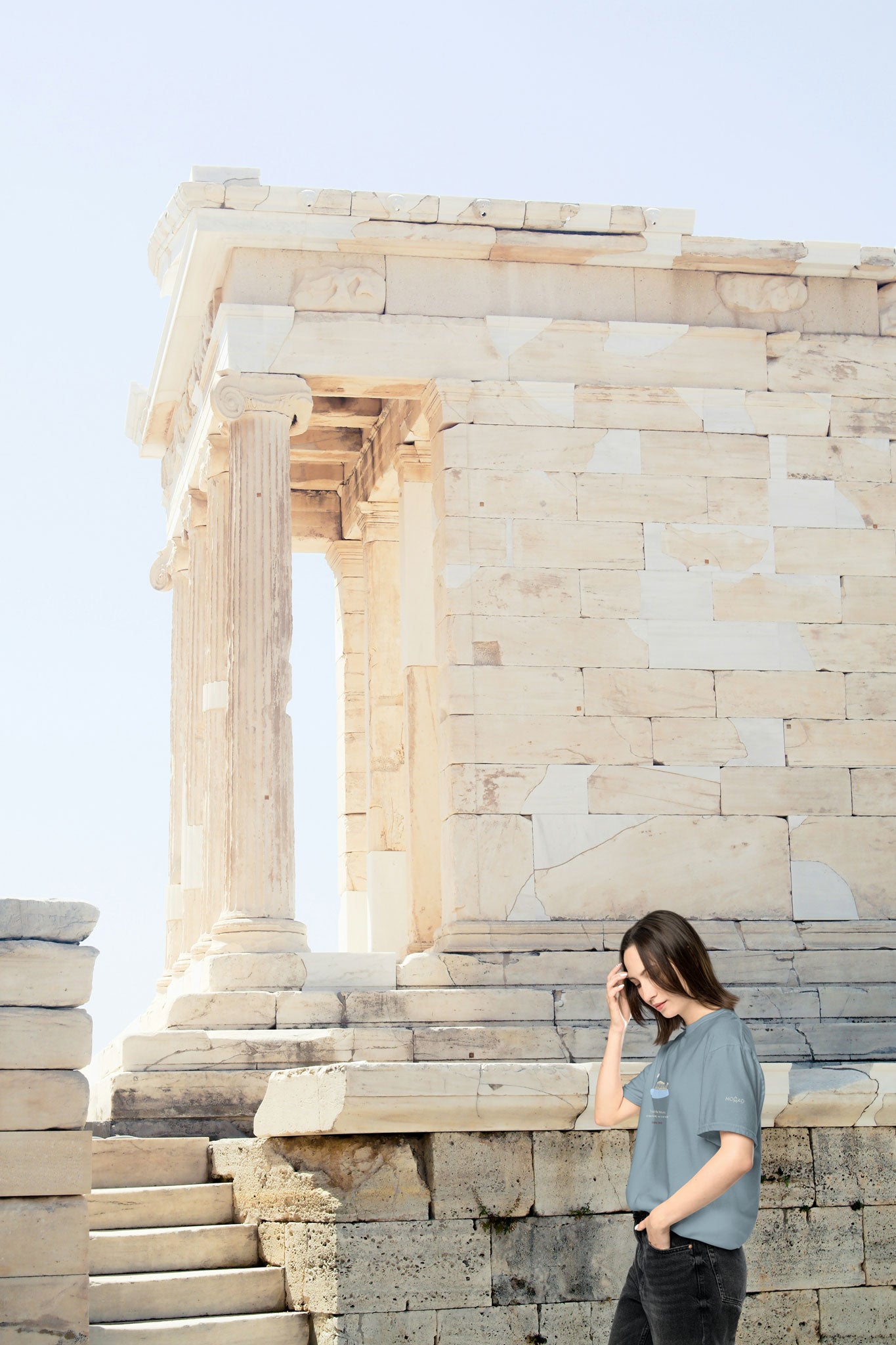 Young woman in ice blue unisex NOMADkit T-shirt with Acropolis Parthenon design, walking near ancient columns in Athens.