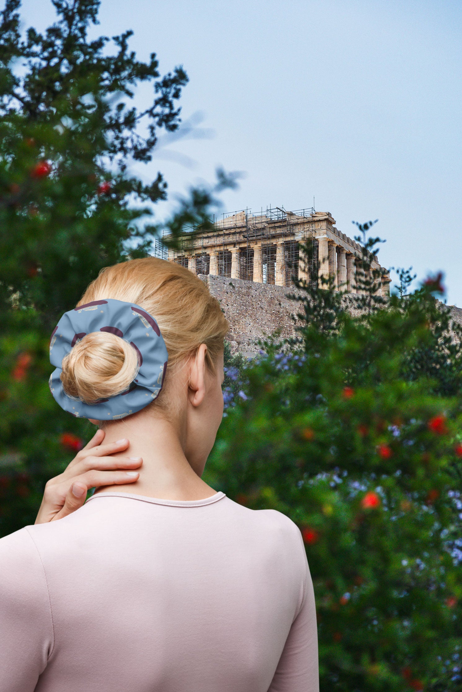 A back view of a woman wearing a NOMADkit scrunchie with an Acropolis design, her hair neatly tied up, while standing in front of the lush greenery with the Parthenon in Athens, Greece, visible in the background.