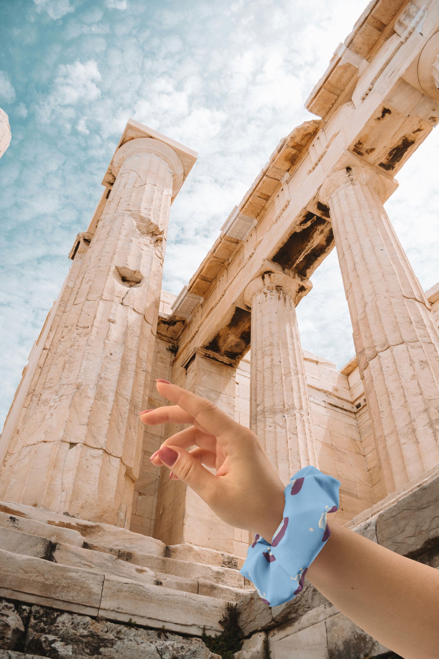 A close-up of a hand wearing a NOMADkit scrunchie with a blue Acropolis design, positioned in front of the historic columns of the Parthenon in Athens, Greece.