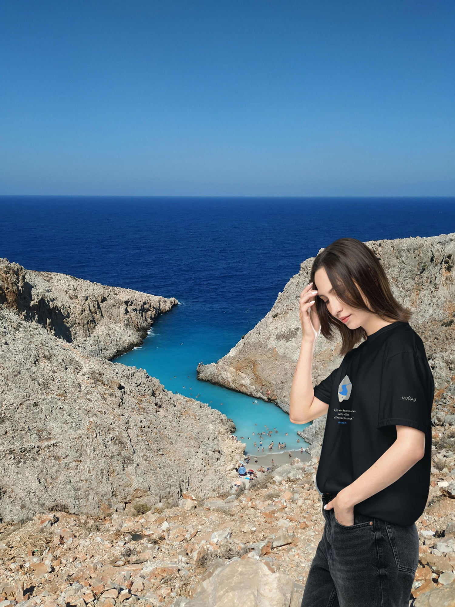 Woman wearing a black NOMADkit t-shirt with Seitan Limania design, standing at Seitan Limania beach in Crete.