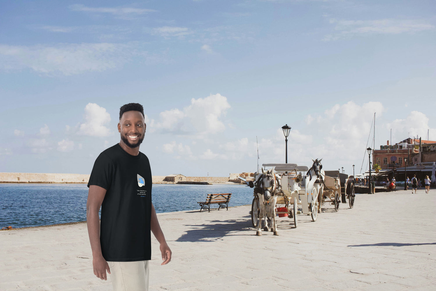 Man wearing a black NOMADkit t-shirt with Seitan Limania design, standing at the Old Port of Chania with horse carriages in the background.