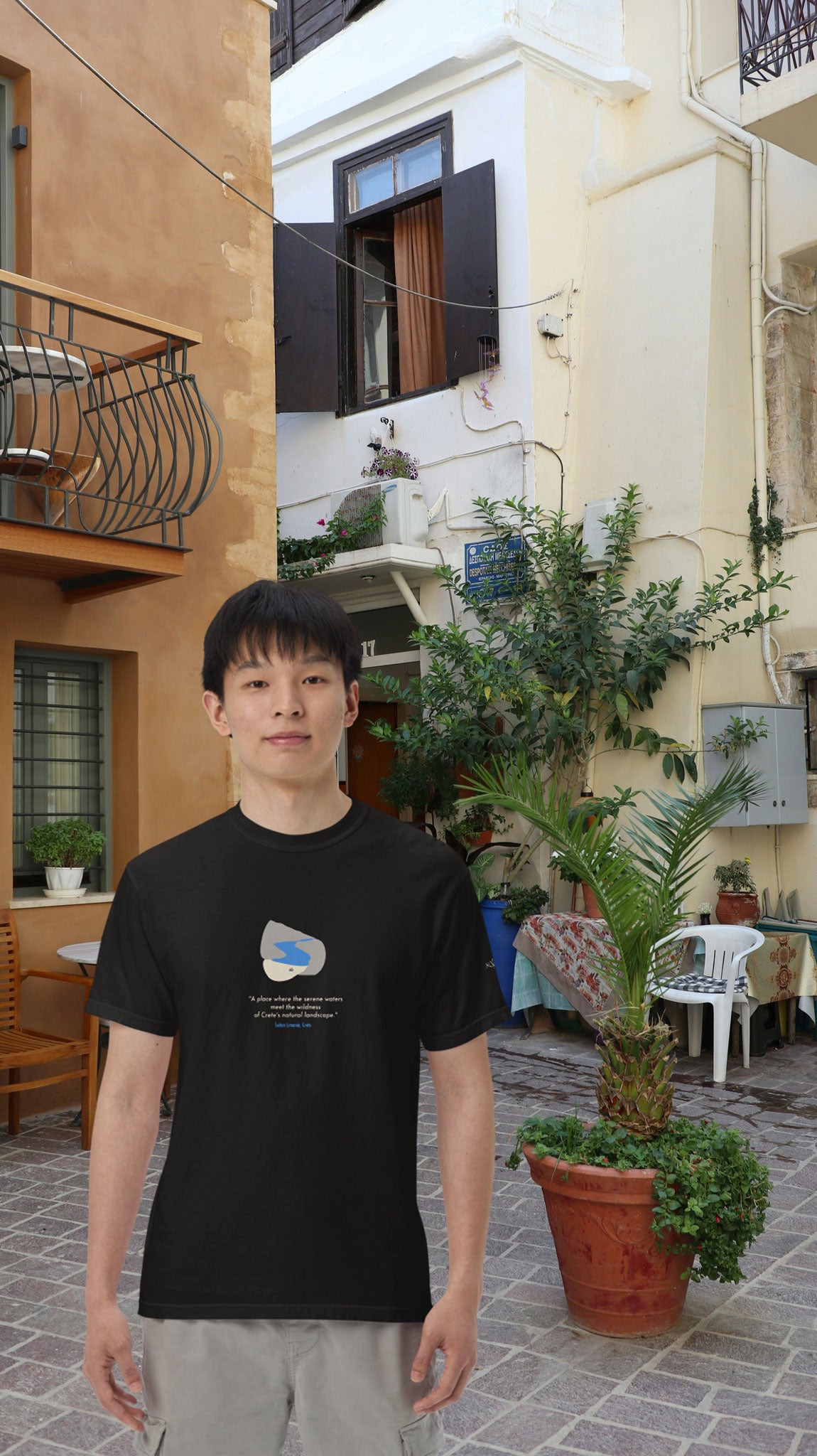 Man wearing a black NOMADkit t-shirt with Seitan Limania design, standing in Chania old town street with traditional buildings.